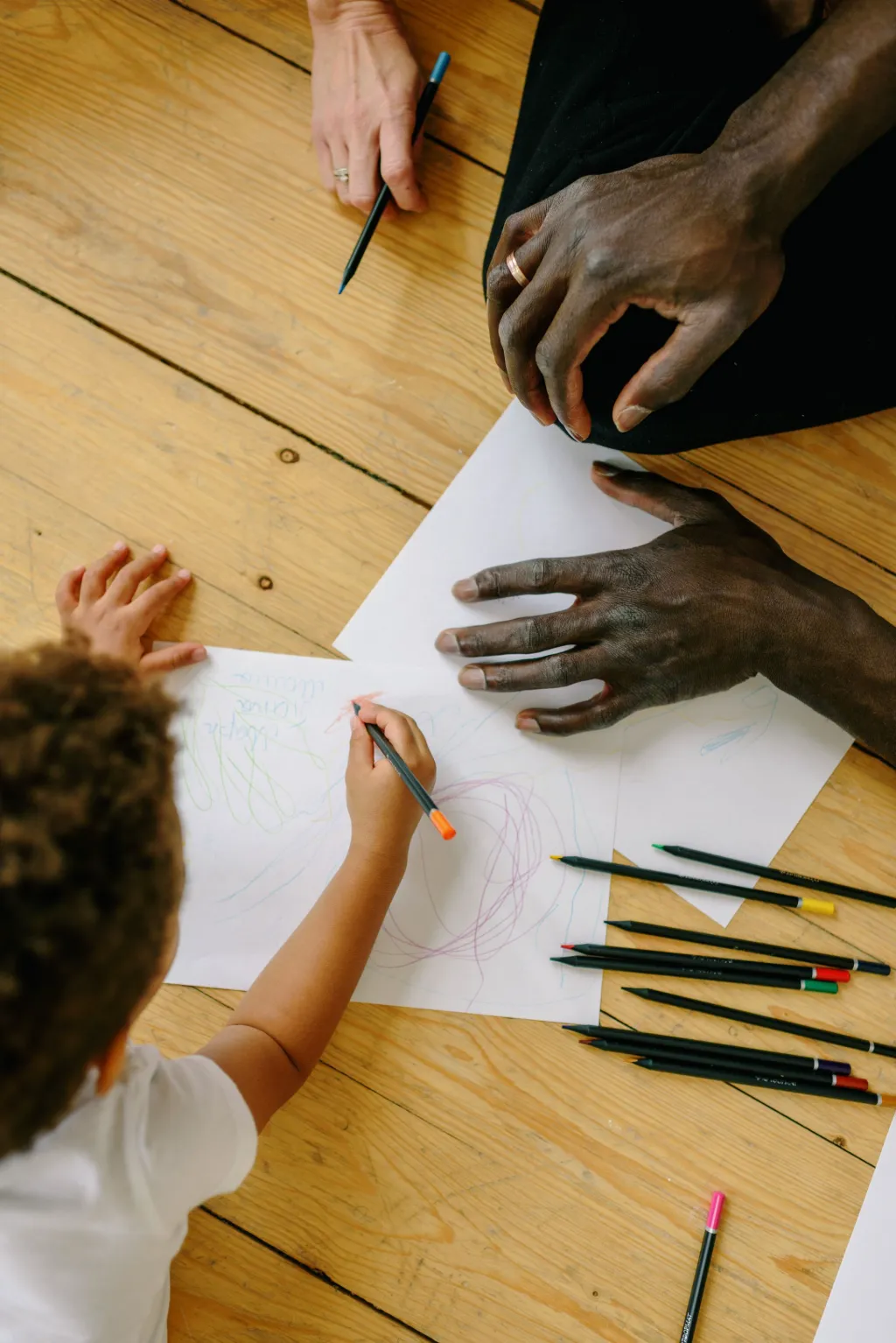 Vu d’en haut, un enfant dessine au crayon à côté des mains de deux adultes